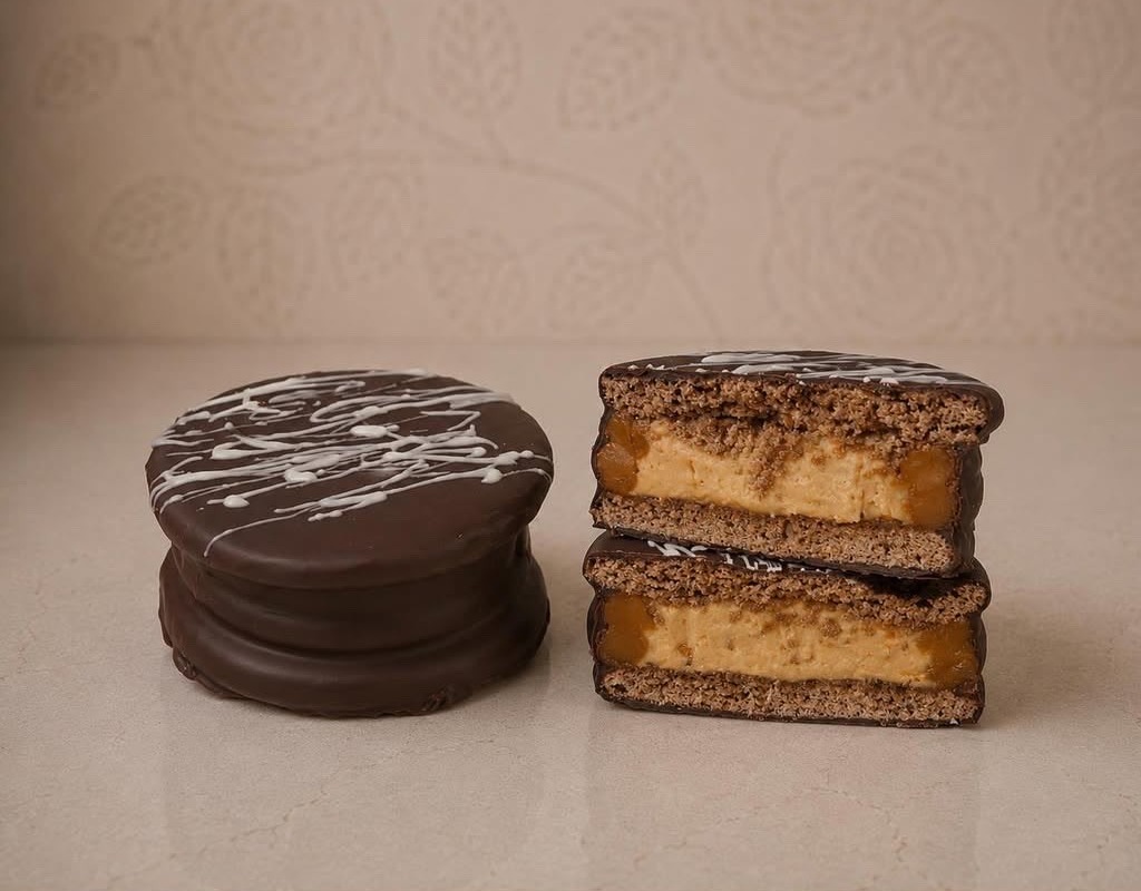 Alfajor bañado en chocolate con hilos de chocolate blanco, entero y en corte mostrando el manjar — Moizlicious San Joaquín